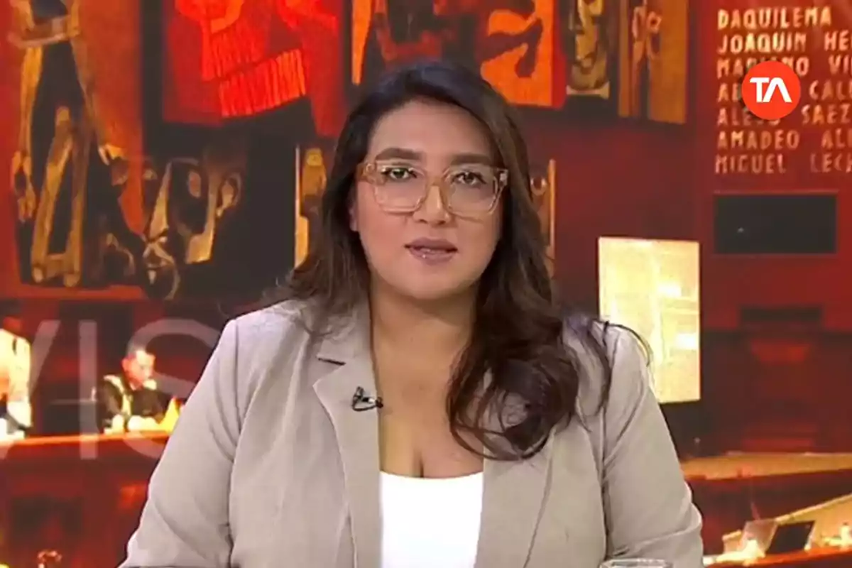 Una presentadora de noticias con gafas y cabello largo está hablando frente a un fondo colorido en un estudio de televisión.