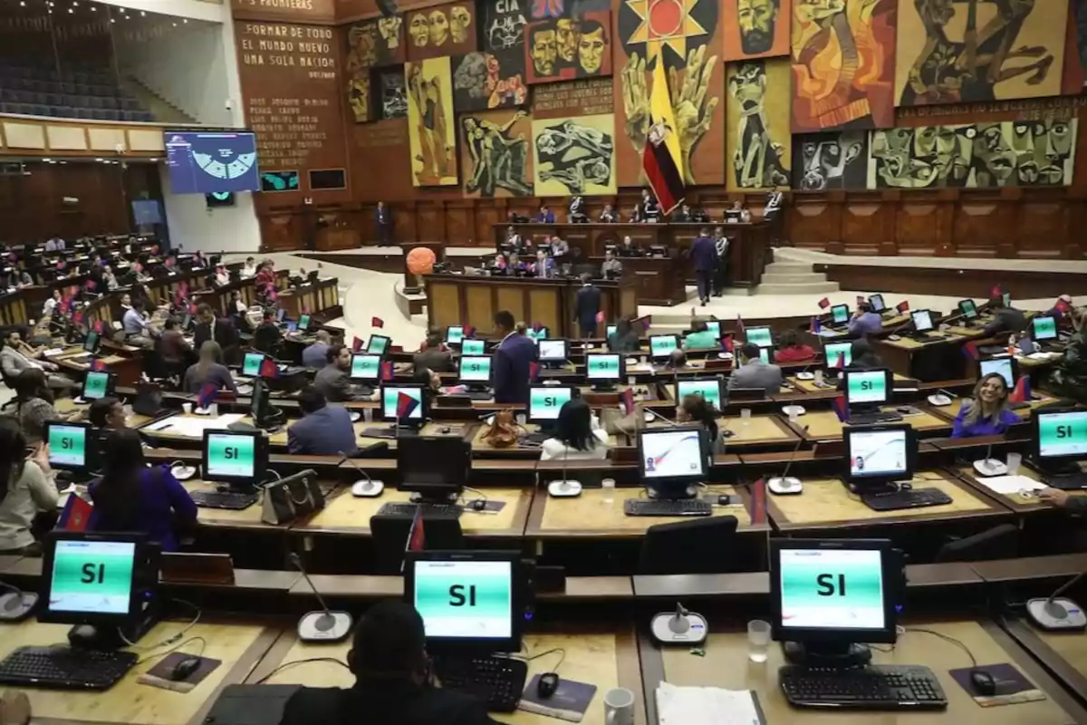 Una sesión parlamentaria con varios legisladores sentados frente a computadoras que muestran la palabra "SI", en un salón decorado con murales y banderas.