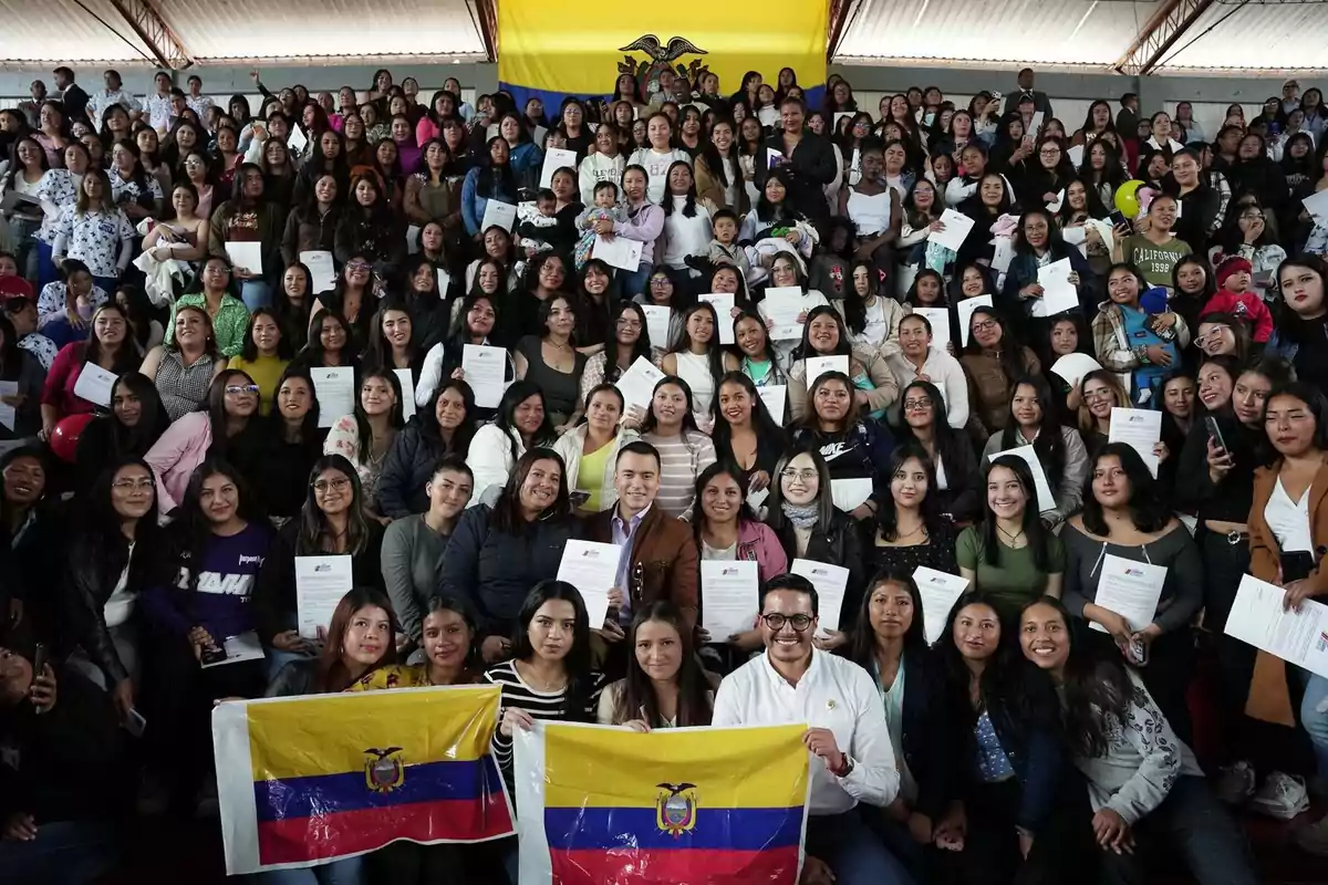 Un grupo numeroso de personas, en su mayoría mujeres, posan juntas sosteniendo certificados y banderas de Ecuador, con una bandera grande del país al fondo.