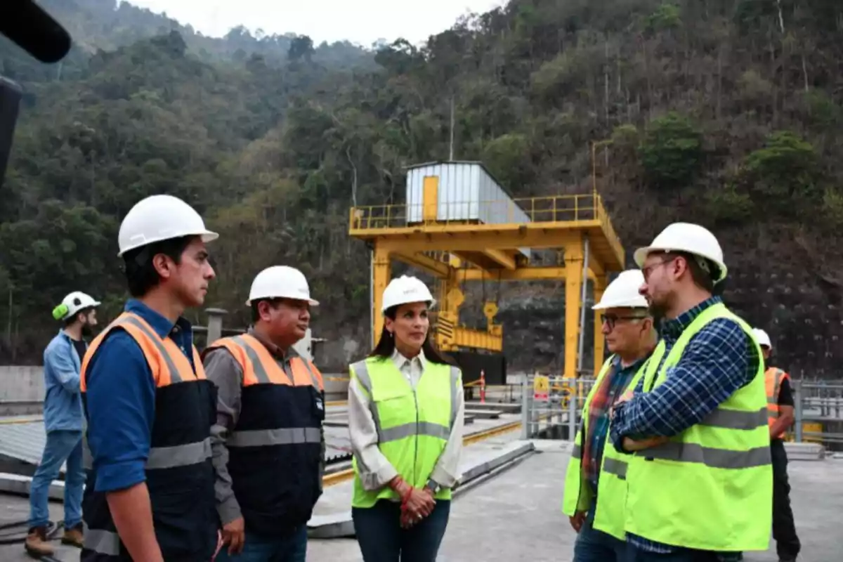 Un grupo de personas con cascos y chalecos de seguridad se encuentra en un sitio de construcción al aire libre con maquinaria amarilla y vegetación en el fondo.