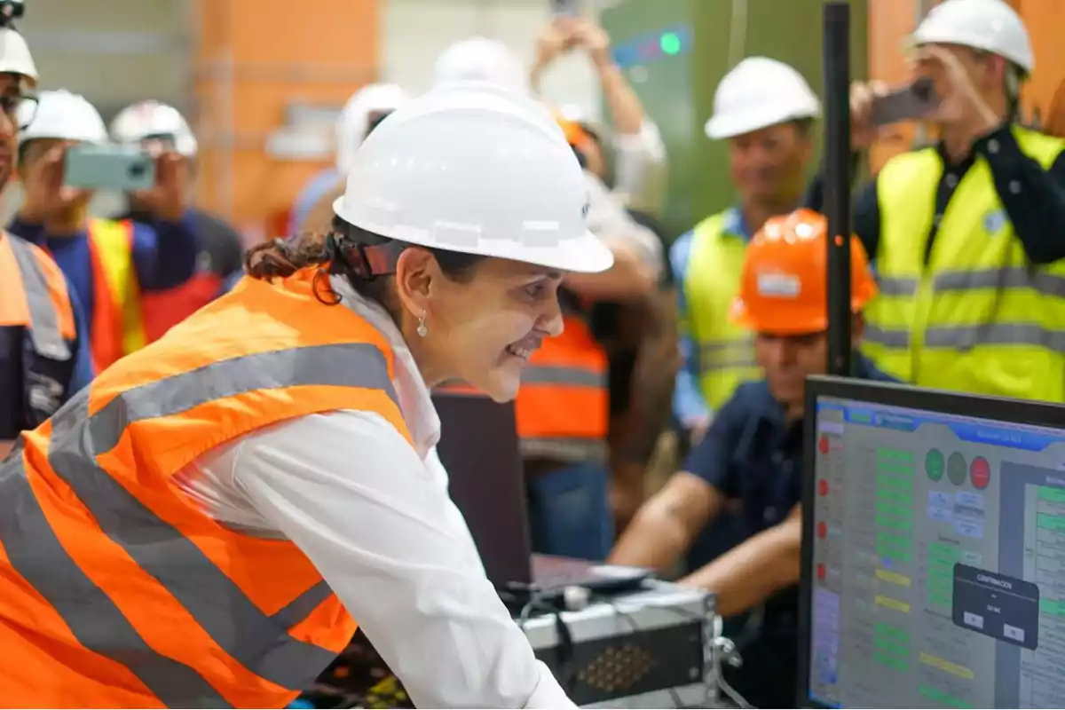 Una mujer con casco y chaleco de seguridad sonríe mientras trabaja en una computadora en un entorno industrial rodeada de otras personas con equipo de protección.