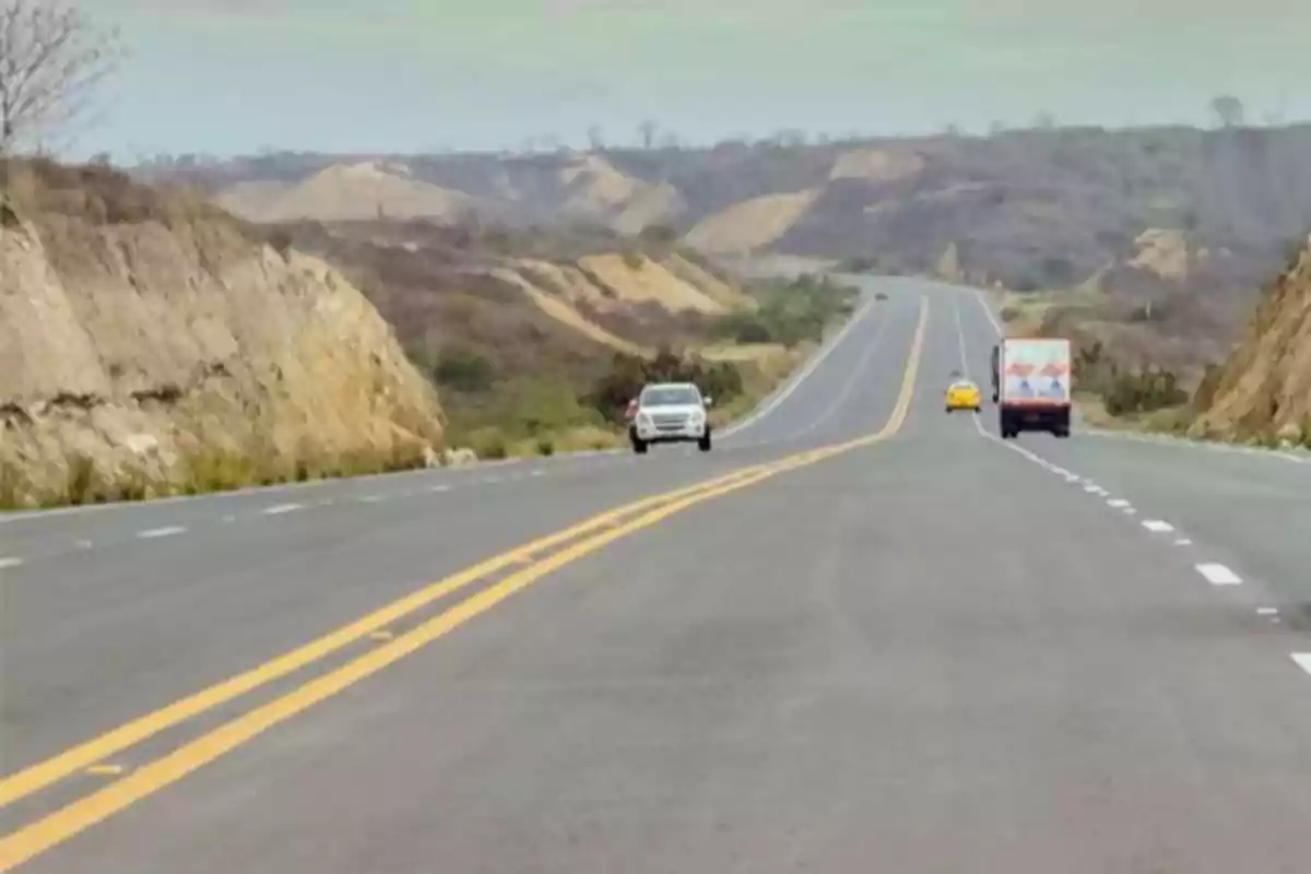 Carretera asfaltada con líneas amarillas y vehículos en movimiento rodeada de colinas áridas y cielo nublado.