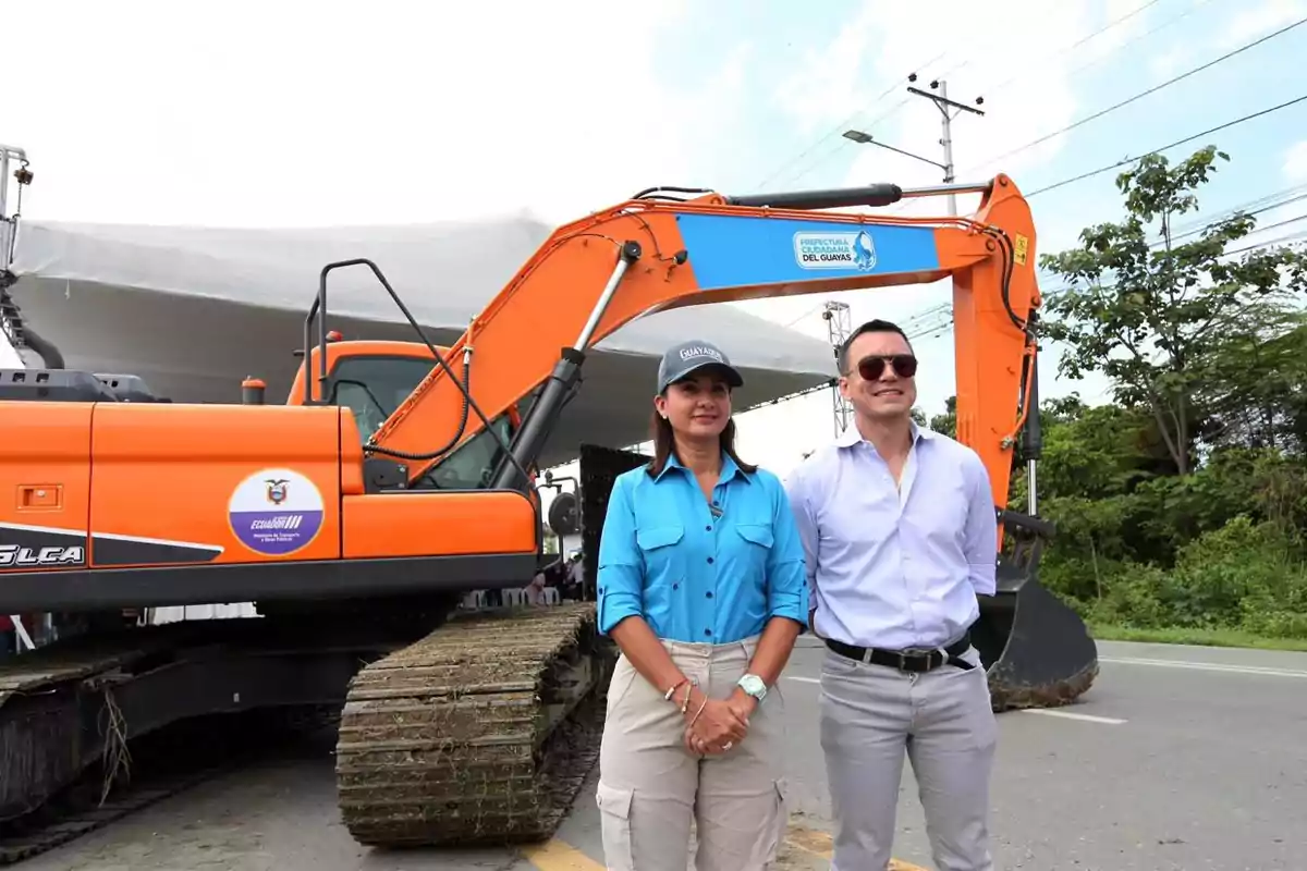 Dos personas posan frente a una excavadora naranja en un evento al aire libre.