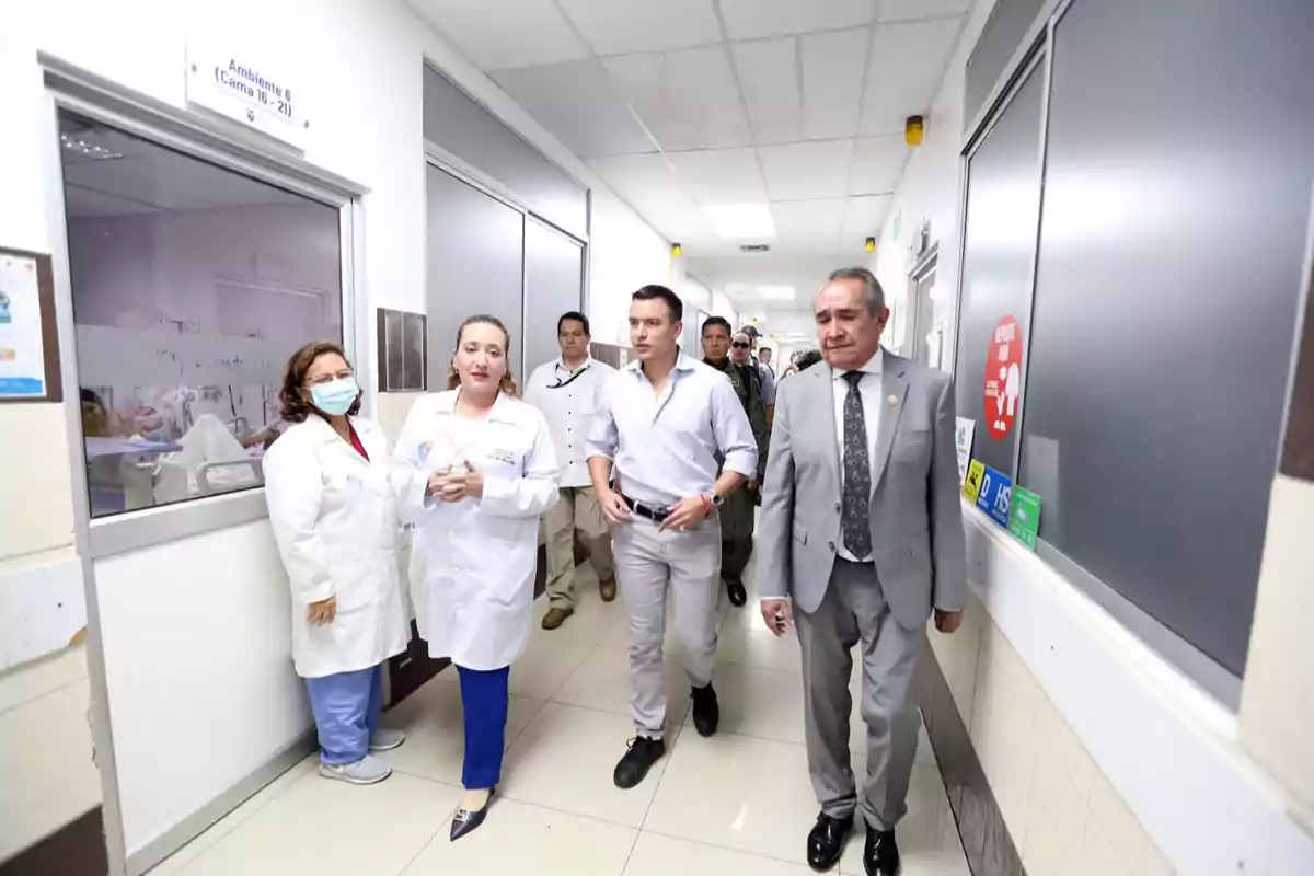 Un grupo de personas camina por un pasillo de hospital, incluyendo personal médico y hombres con traje.