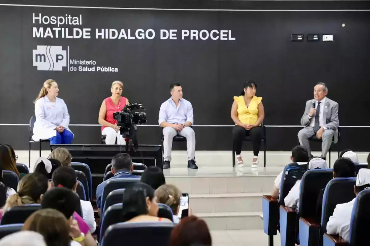 Un grupo de personas sentadas en un escenario frente a una audiencia en el Hospital Matilde Hidalgo de Procel, con un cartel del Ministerio de Salud Pública en el fondo.