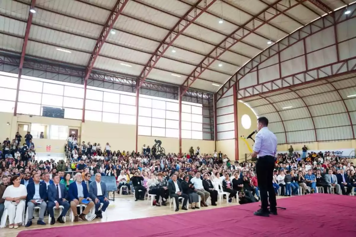 Una persona hablando en un micrófono frente a una gran audiencia en un gimnasio techado.