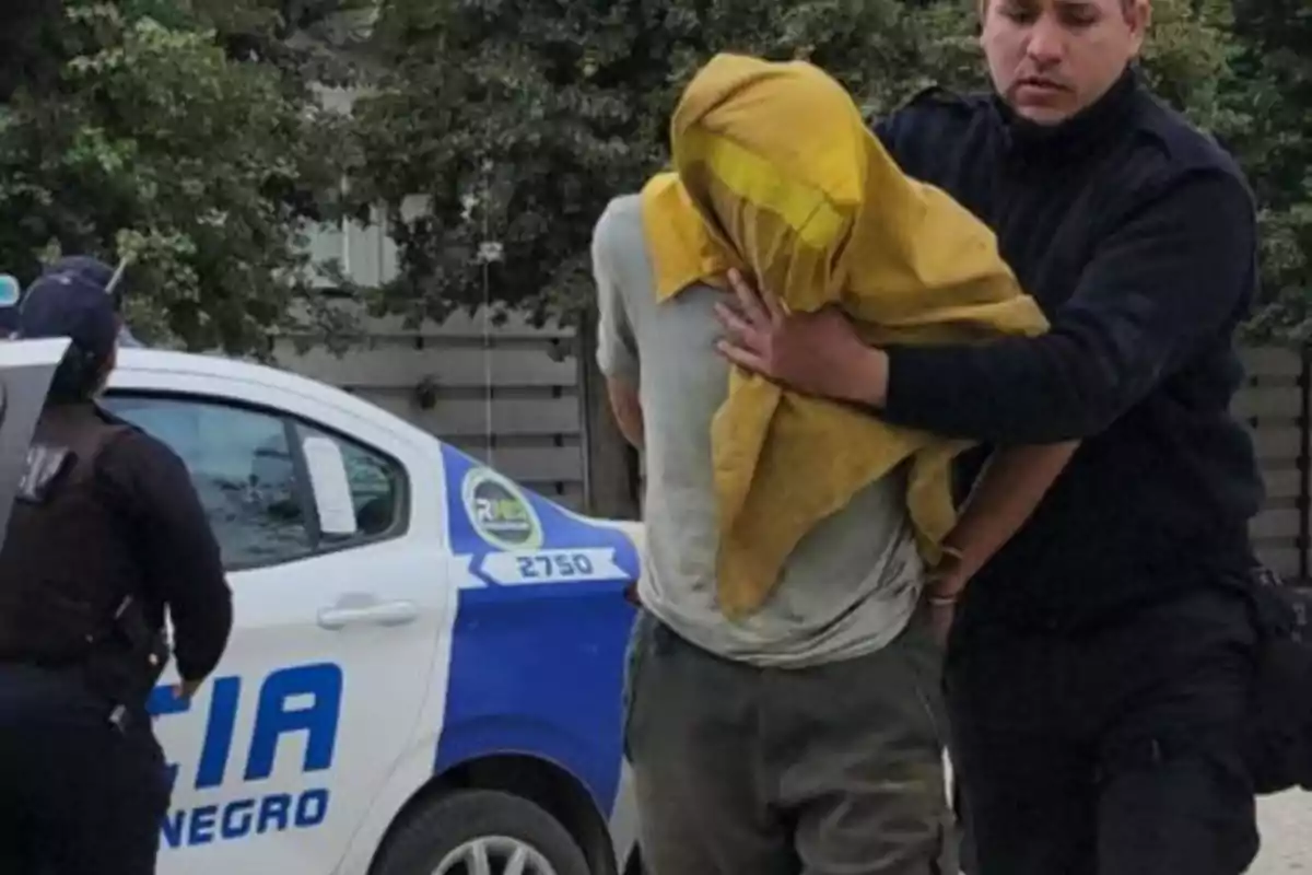 Un policía detiene a una persona con la cabeza cubierta junto a un coche patrulla.