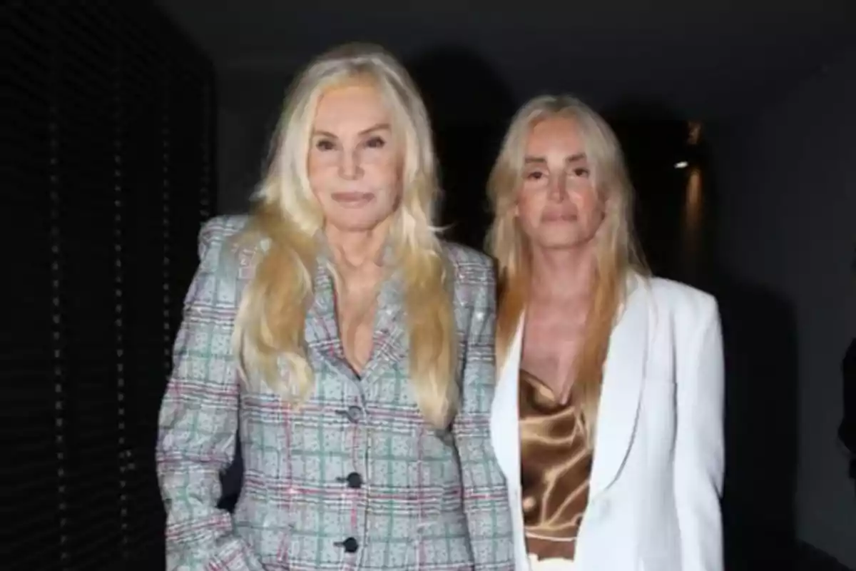 Two women with blonde hair pose together, one wearing a checkered suit and the other a white outfit with a gold blouse.