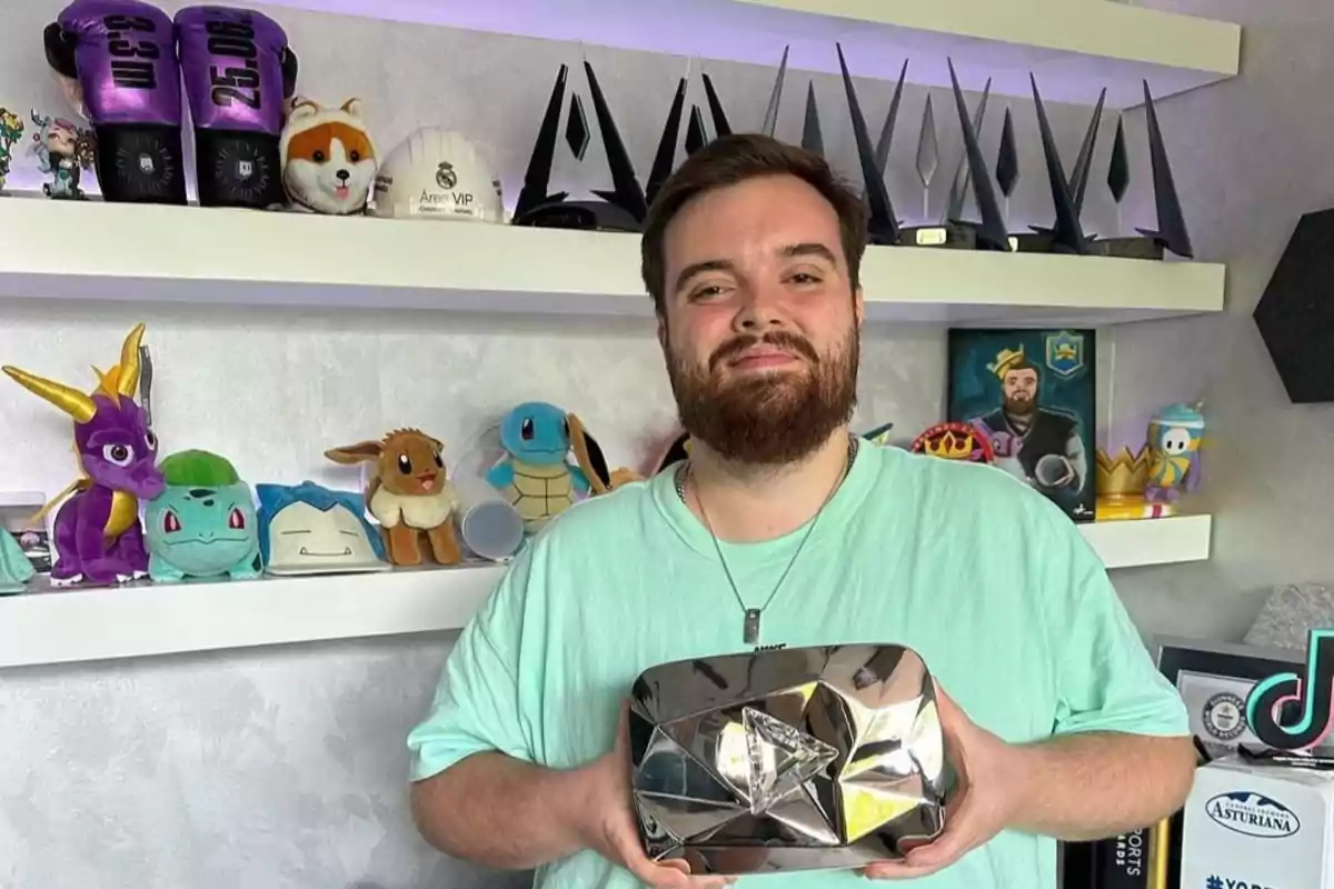 A man holds a YouTube Diamond Play Button in front of a shelf full of stuffed animals and trophies.