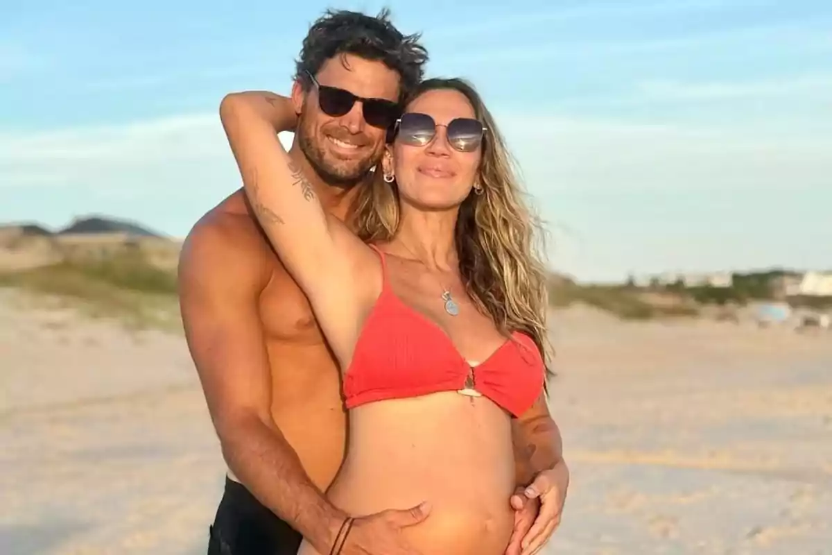 A smiling couple on the beach, with the pregnant woman and the man hugging her from behind.