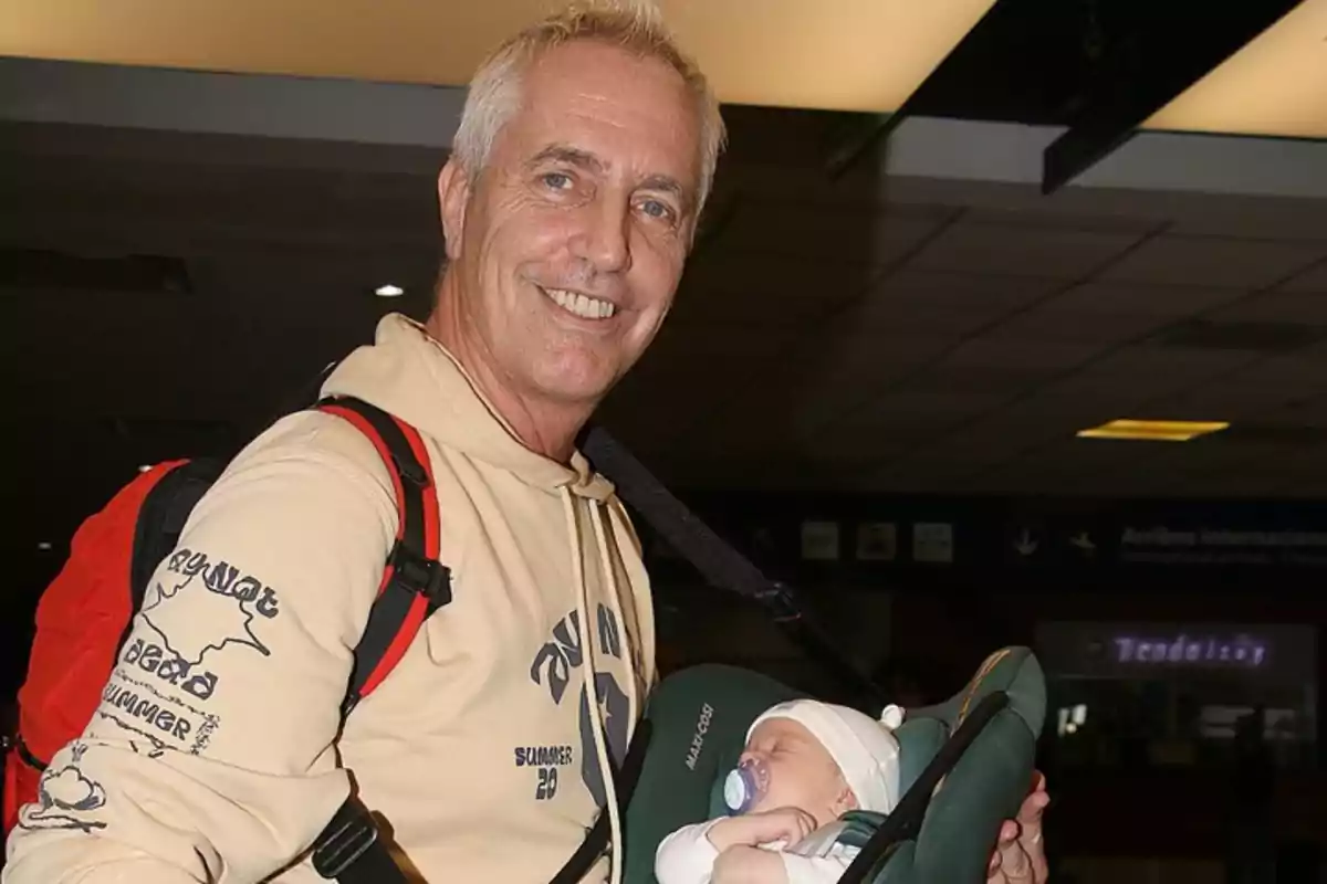 A smiling man carries a sleeping baby in a baby carrier in a public place.