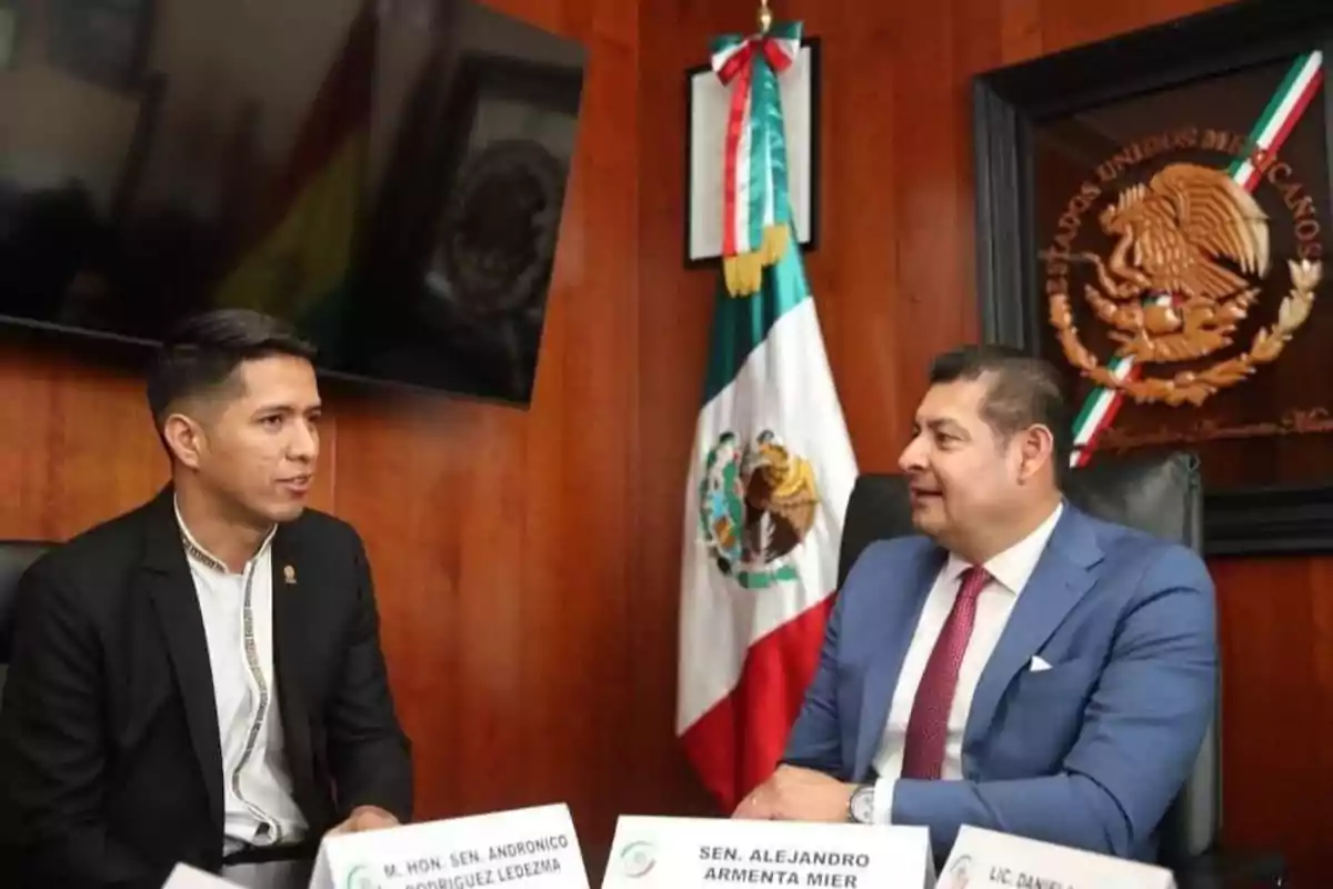 Two men in suits are talking in an office with a Mexican flag and a coat of arms on the wall.