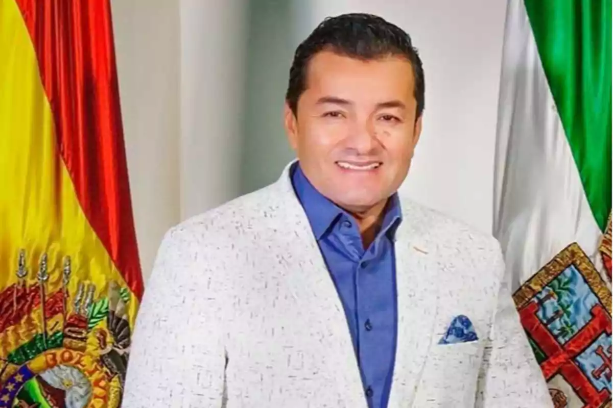 A man in a white jacket and blue jersey is standing in front of the flags of Bolivia and Mexico.