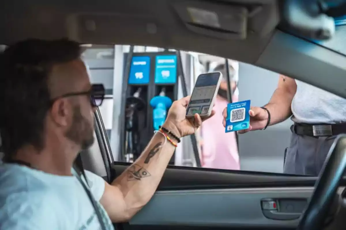 Un hombre en un auto sostiene un teléfono mientras otra persona le muestra un código QR en una estación de servicio.