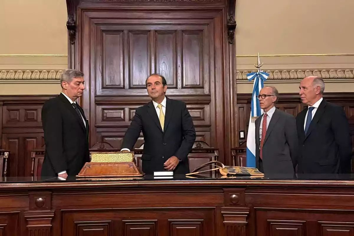 Un grupo de hombres en traje formal se encuentra en una sala con un fondo de madera tallada y una bandera azul y blanca.