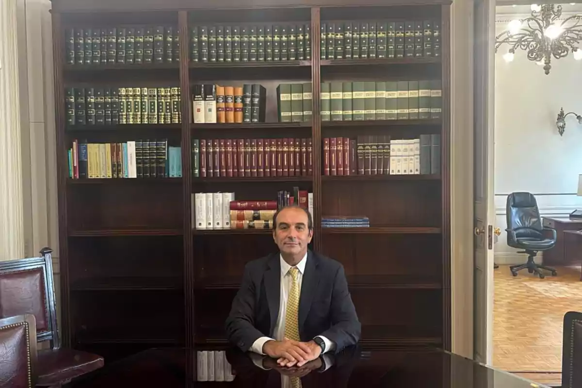 Un hombre sentado frente a una mesa en una oficina con estanterías llenas de libros detrás.