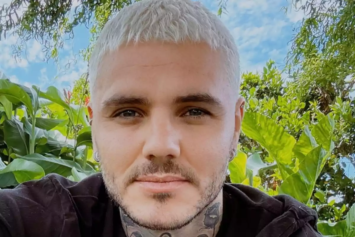 Un hombre con cabello corto y rubio platino, barba y tatuajes en el cuello, posando al aire libre con plantas verdes y cielo azul de fondo.