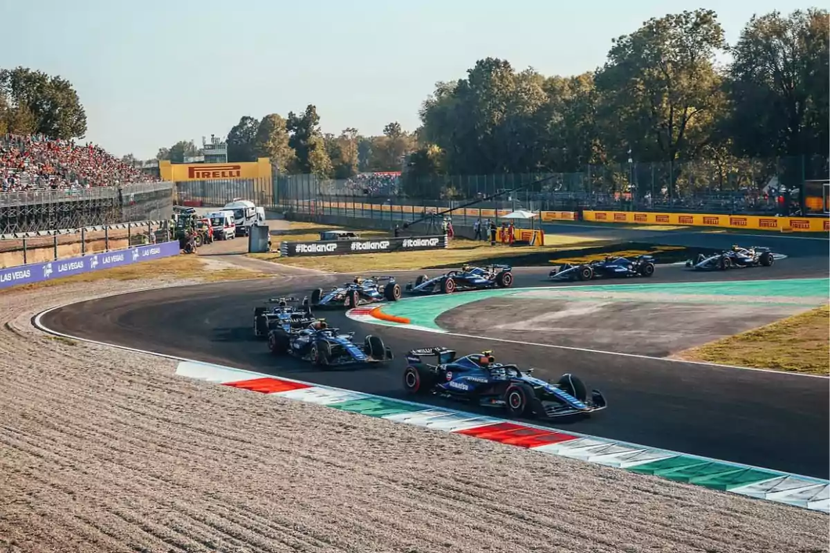 Carrera de autos de Fórmula 1 en una curva del circuito con público en las gradas y árboles al fondo.