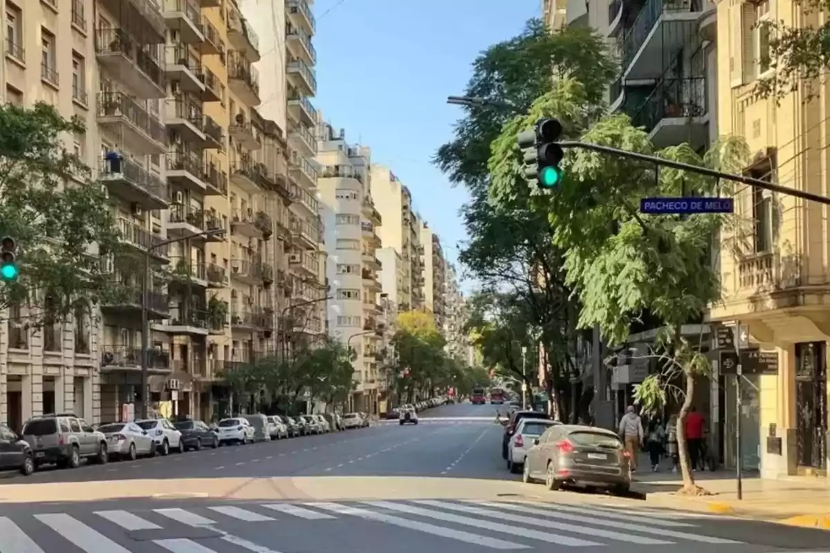 Calle urbana con edificios altos a ambos lados, árboles y coches estacionados, un semáforo en verde y una señal que indica "Pacheco de Melo".