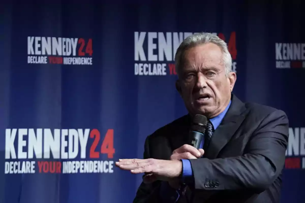 A man speaking at an event with a microphone in hand, in front of a background that says 