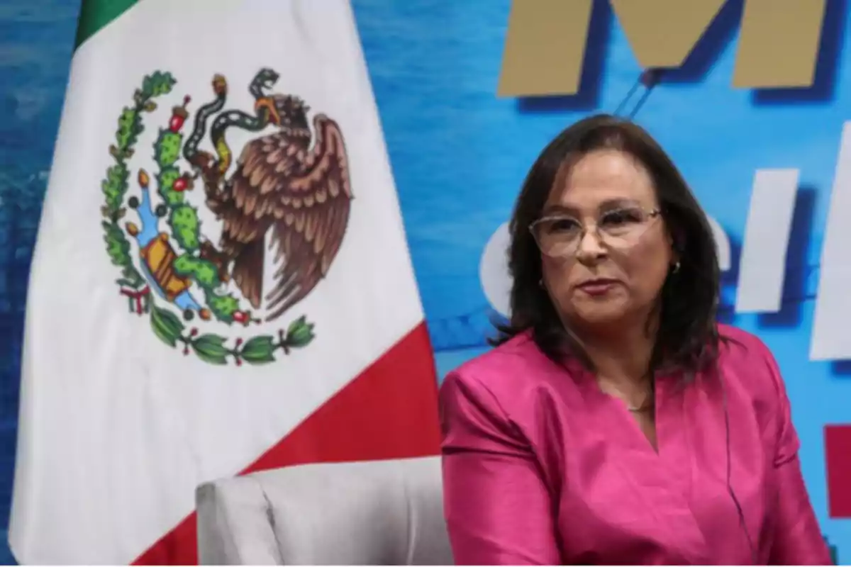 A person with glasses and a pink jacket is sitting next to a Mexican flag.