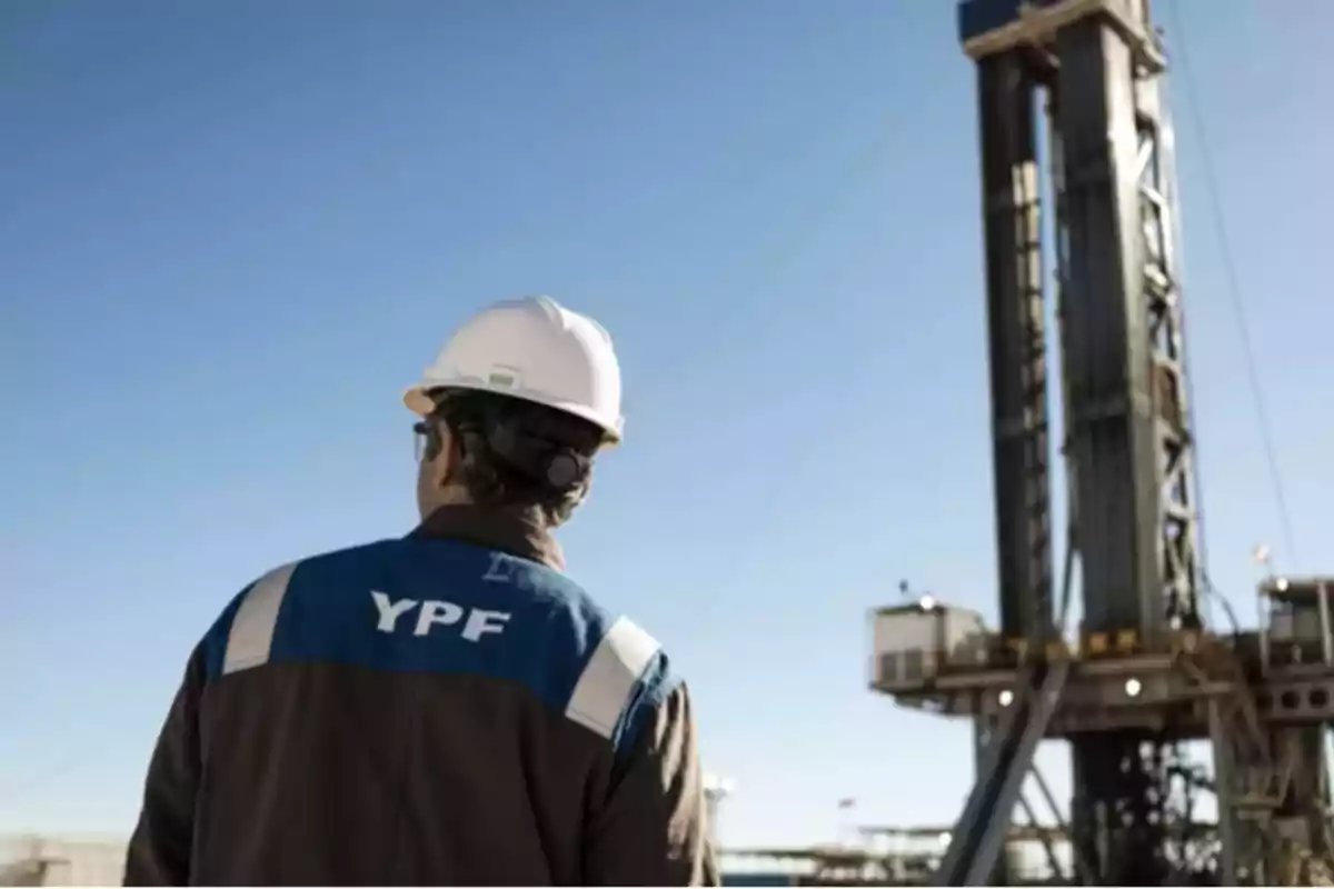 Persona con casco de seguridad observando una torre de perforación en un campo petrolero.