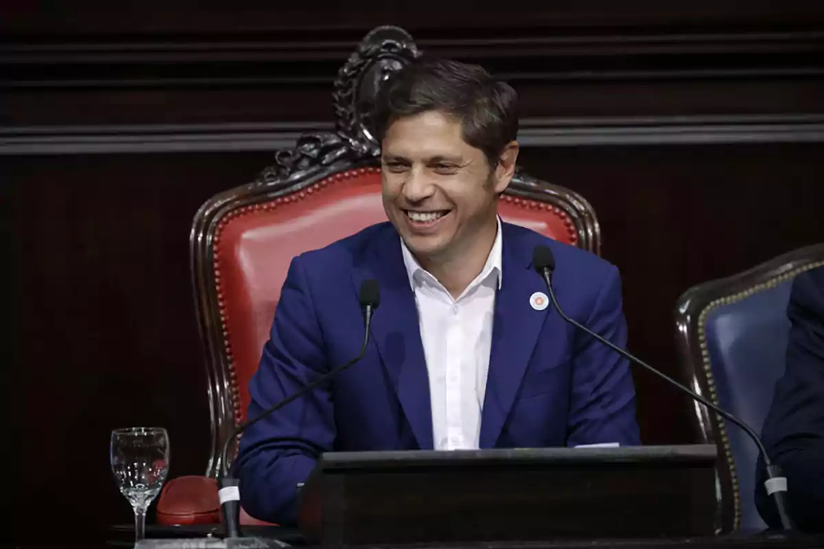 Un hombre sonriente con traje azul sentado en una silla de respaldo alto en un entorno formal.