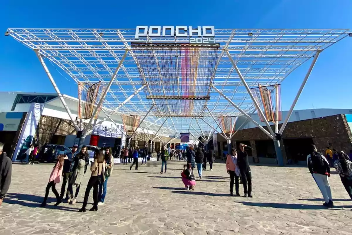 Personas caminando bajo una estructura metálica con un cartel que dice "Poncho 2024" en un día soleado.