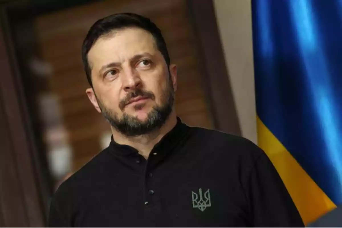 Hombre con barba y cabello corto, vistiendo una camiseta negra con un emblema, de pie junto a una bandera azul y amarilla.